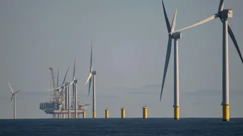 Dogger Bank Photo of large offshore white wind turbines with the sky in the background. There are about six wind turbines in a row with a ship in the background also. The turbines are spinning. There are some clouds in the sky. 