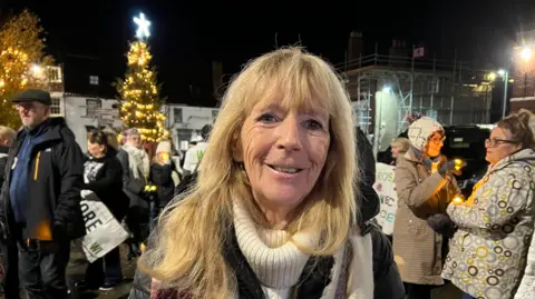 BBC/Becki Bowden Woman with blonde hair and white jumper wearing a dark coat. There is a crowd behind her.