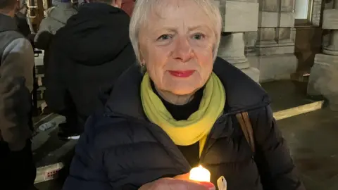 BBC/Lara King Woman with short white hair, green scarf and black jacket. She is holding a candle. She is stood on a street.