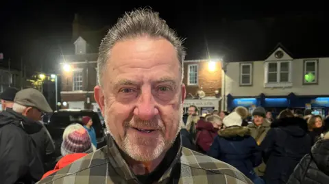 BBC/Becki Bowden Man with short hair and short beard with chequered jacket. There is a crowd behind him.