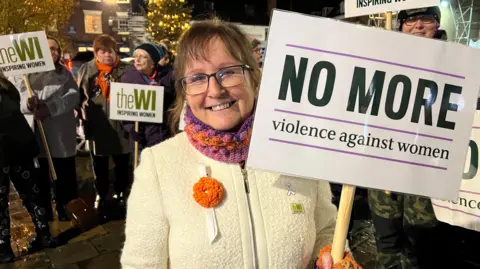 BBC/Becki Bowden Woman with brown hair, white jumper, scarf, she is wearing glasses and holding a placard which reads, no more violence against women. There is a crowd behind her.