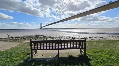 Shropshire Liam An image of the Humber Bridge against a blue sky dotted with clouds. In the foreground is a wooden bench casting a long shadow over the grass. 