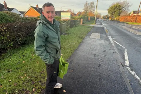 A man a green jacket and black trousers is carrying a shopping bag. On the pavement next to him are a series of footsteps moulded into the tar, which have since set.