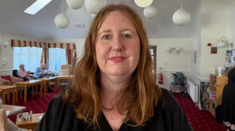 The picture shows a woman, Natalie Spikings, smiling. She has shoulder-length red hair, and is wearing a black top. She is standing in front of a room which has a red carpet and lots of tables and chairs. There are two elderly women chatting behind her. 