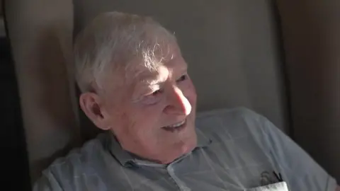 Fred Holmes sat in a beige chair smiling, looking to the right of the camera. He is wearing a grey collared short-sleeve shirt with large checks on. He has short grey hair.
