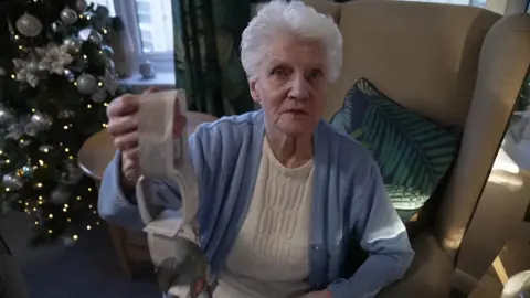 BBC Jean Waddy sits in a beige armchair. She has short grey hair and is wearing a white top and a light blue buttoned sweater. She is looking at the camera and holding up a paperchain decoration made out of newspaper. A Christmas tree covered with silver decorations is lit up on the left.