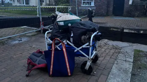 A trolley full of bags of belongings sitting on a brick path next to a canal. Behind it is a meal fence with black and yellow tape