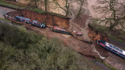 PA Media An aerial view of the collapse - showing water that has run into a nearby field. There is a large hole in a canal and the section is dry with no water. Two boats are in the hole, with another on higher dry ground hovering over the edge into the hole