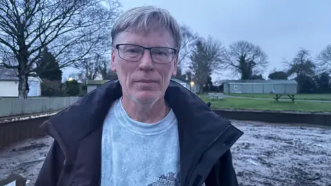 A man with grey hair and black glasses standing with a dirty canal behind him