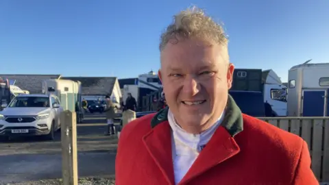 Jake Zuckerman/ BBC Gerrard Farmer stood smiling at the camera wearing a red coat and a white shirt. He has short light brown hair. People on horses can be seen behind him. The sky is blue. 