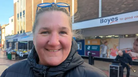 A woman with blond hair, tied back, smiles as she looks at the camera. She has blue-framed glasses on top of her head and is wearing a black winter coat. She is standing in a pedestrian high street in front of a store with a large white sign reading "Boyes" in blue letters and "for good value" in red letters.