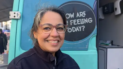 A woman with dark grey hair wears a navy-blue fleece and round, brown-framed glasses as she stands in front of the back doors of a blue van. A logo on the van, partially obscured, reads: "How are you feeling today?". Within the van can be seen a first aid kit and a worksurface with some medical equipment on it.