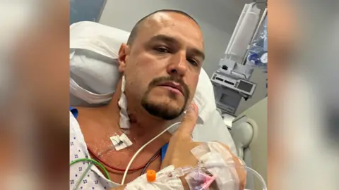 Reece Holland A man with shaved brown hair and a goatee beard give the thumbs up as he lies in a hospital bed. He is connected to has numerous tubes and wires connected to him as well as a dressing on the side of his neck. He is giving a thumbs up to the camera.