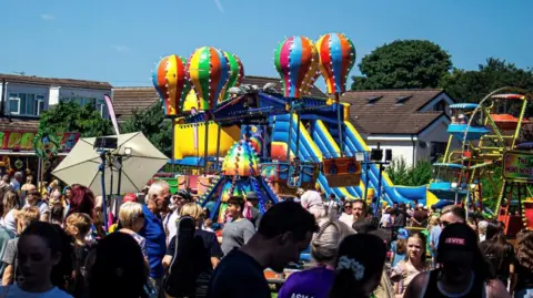 Ann Foster A lively outdoor carnival scene on a sunny day, featuring colourful amusement rides such as hot air balloon-themed attractions, inflatable slides, and a Ferris wheel. The area is crowded with people enjoying the event, surrounded by vibrant stalls and signage.