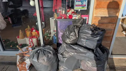 Dove House Hospice Charity Shop Black bags piled on top of each other at the entrance to Dove House Charity shop. The store's window display includes Christmas presents and teddy bears. 
