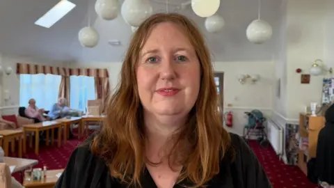The picture shows a woman, Natalie Spikings, smiling. She has shoulder-length red hair, and is wearing a black top. She is standing in front of a room which has a red carpet and lots of tables and chairs. There are two elderly women chatting behind her.
