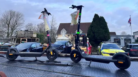 Two electric scooters, both black with colourful flashes, stand on a metal platform in a yard under a grey-blue sky. They have seizure notices attached to the handlebars. Various buildings can be seen in the background, including a house, a small grey portable building and a shed-type structure. To the right, a Union flag flies on a white flagpole.