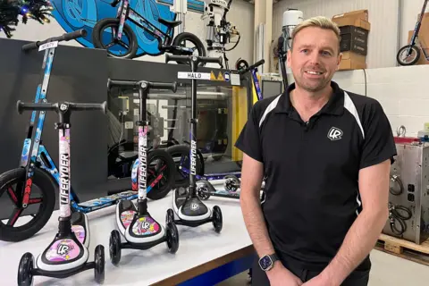 A man with short blonde hair, slicked back, and stubble smiles as he stands in a workshop in front of a number of scooters of various sizes on a white shelf. He is wearing a black polo shirt. The scooters have colourful liveries and logos reading "Liferyder". 