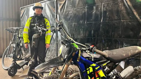 BBC A female police officer dressed in a black uniform, yellow hi-vis vest and white hat stands in a warehouse surrounded by electric bikes and scooters. Behind her is a black screen. The bike closest to the camera resembles a scrambler motorcycle, with a large saddle and colourful cowling.