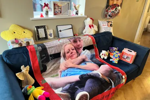 A blue sofa covered in the contents of a Christmas Eve box - there is a blanket with a mother and son on it, teddies, sweets, frames, certificates and other keepsakes.
