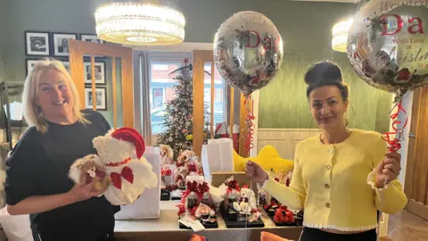 BBC Two women stand by a table full of gifts for Christmas Eve gift boxes, including teddy bears, balloons and blankets. The woman on the left has bobbed blonde hair and a black t-shirt. The woman on the right has dark hair in a bun and wears a yellow cardigan, they are both smiling. 