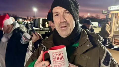 Phillip Norton/BBC A man with a woolly beanie hat and brown jacket looking towards the camera. He is holding a red and cream-coloured mug which has the word "Annie" on it. There is another man wearing a Santa hat on in the background.