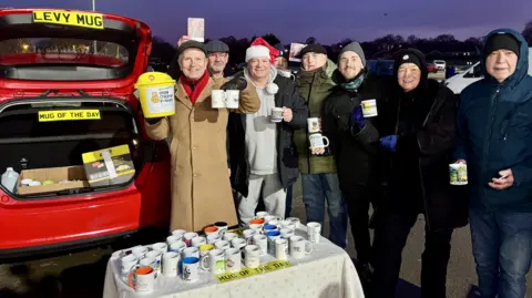 Phillip Norton/BBC Look North presenter Peter Levy pictured during his mug giveaway in Hull. He is wearing a long brown coat and green cap and is standing next to a red Ford Fiesta with an array of mugs on a table in front of the vehicle. There are several other people in the shot holding mugs.
