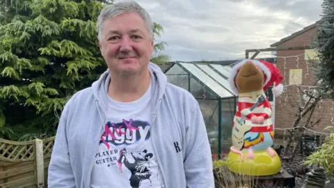 Amber Macey/BBC Jon Seth - a man wearing a grey hoodie and rat t-shirt honouring Mick Ronson - standing in front of a giant red and white striped rat sculpture holding a guitar. The sculpture has a red Santa hat on its head.