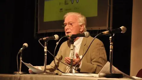 Doc Rowe A white haired man in a beige jacket sits at a table behind several microphones.