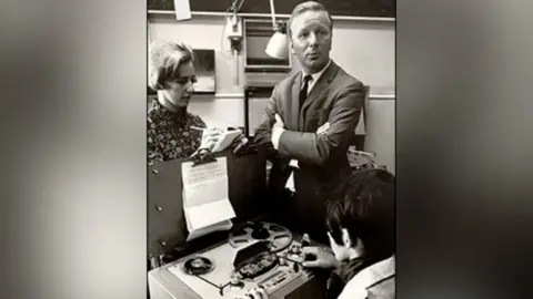 BBC A sharply dressed man in a suit stands with his arms folded whilst a woman takes notes onto a a pad of paper. Another women sits in-front of an old fashioned tape reel player