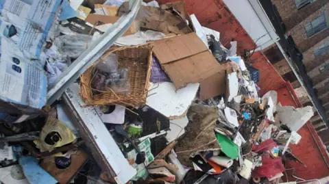 Maxine Rolling A pile of household belongings in a skip.