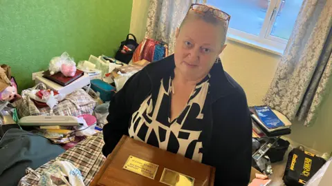 Amanda White/ BBC Maxine Rolling stood in a room full of salvaged items, holding a brown box that contains her dad's ashes. She is wearing a black and white V-neck blouse and a black zip-up fleece. Her light brown hair is tied back and she has tortoise-shell glasses on her head. The room she is stood in has green walls, a window behind her and patterned curtains. 