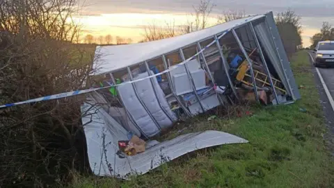 Maxine Rolling A large white lorry full of belongings tipped on its side on a roadside grass verge. 