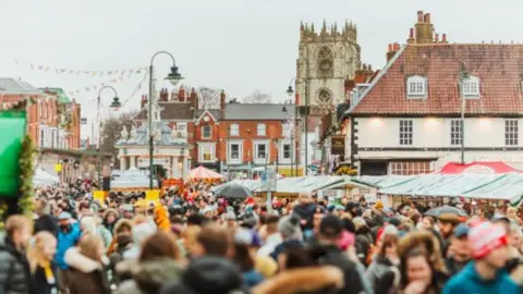 East Riding of Yorkshire Council Crowds of people dressed in winter clothing, including some wearing hats, walk past a range of stalls, with a church and other historic buildings in the background. 