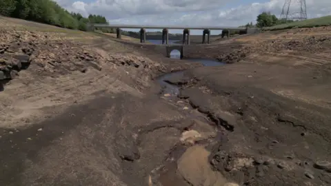 Spencer Stokes/BBC A bare reservoir bed with a bridge in the background