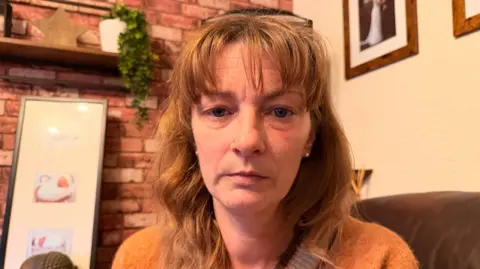 BBC News/Jasmine Lowe A lady with ginger hair and a fringe, she has black glasses on her head and is sat in a living room. The lady has pale skin and blue eyes and is wearing an orange jumper. 