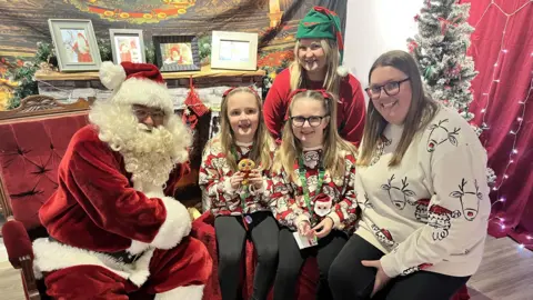 BBC Father Christmas, two twin girls wearing matching Christmas jumpers, their mother, also in a Christmas jumper, and a female elf assistant, all pose for a photo in a Santa's grotto. They are all smiling. 
