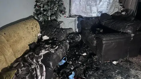 Jasmine Lowe/BBC News A living room completely smoke damaged, it has ash everywhere and most items have burned down. The Christmas tree especially has completely burned. 