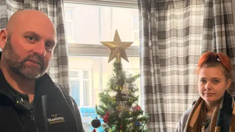 BBC News/Jasmine Lowe A bald man, wearing a jacket stood on the right of a fake decorated Christmas tree, next to him is his daughter who has red hair in a bun wearing a scarf. 