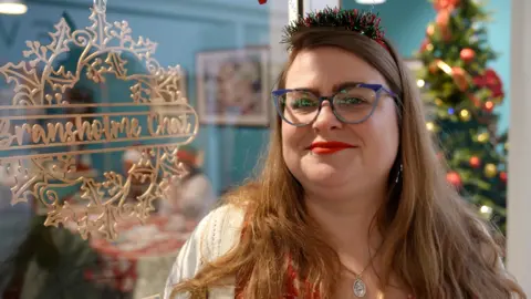 A woman wearing a festive tinsel headband, blue-rimmed glasses and red polka-dot top stands near a glass door with a frosted “Bransholme Chat” design on it. She wears bright red lipstick, a silver necklace and a beige cardigan. There is a decorated Christmas tree in the background. 