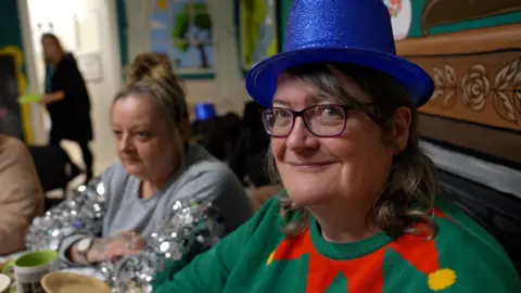 BBC Two women are seated at a table during a festive gathering. Carrie in the foreground, wears a bright blue glittery top hat and a green jumper with a red and yellow Christmas design. The table is set with cups, plates, and silver tinsel decorations, and a colorful mural is visible in the background. The second woman has blonde hair in a bun and is wearing a grey and silver sparkly top.