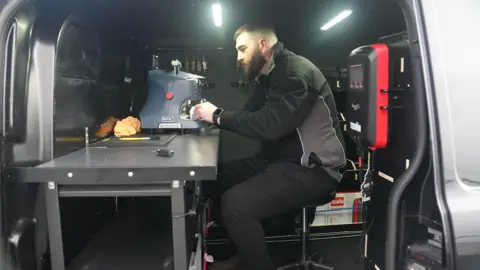 Sam is sat in the back of his van at a desk cutting a key with a machine.