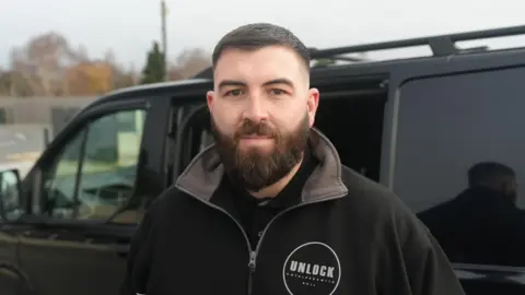 BBC A man with dark hair and a beard looks straight to the camera. He is wearing a black jacket with the name of his company Unlock on the front