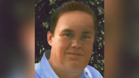Missing People Image of how Philip Fudge, who went missing at the age of 21 in 1995, could look today. He has brown hair and is wearing a blue shirt. He is in front of some shrubs. He is smiling.