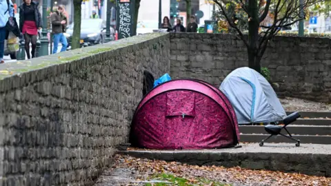 EPA A pink and a blue tent are pitched on concrete steps. On the other side of the wall people walk past carrying shopping bags.