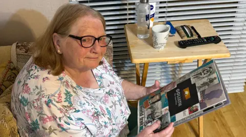 Kevin Shoesmith/BBC News A bespectacled woman with greying light brown hair holds a scrapbook with pictures of her son pasted on to the cover.