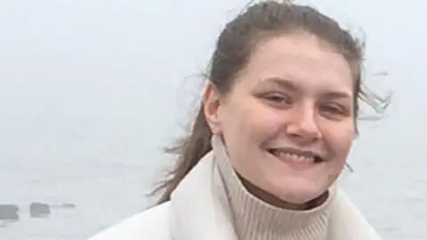 Squire family Family picture of Libby Squire. She has long dark hair and is wearing a white jumper and coat