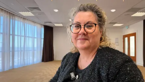 Jasmine Lowe/BBC A picture of Lisa Squire. She has short blond hair and wears glasses and a black top. She is standing in an empty room and looking into the camera.