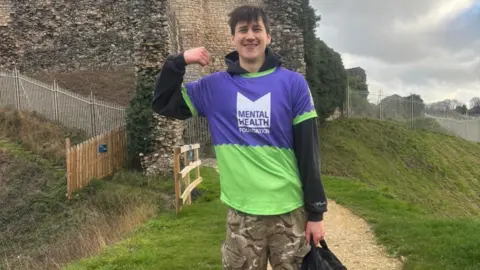 Supplied A young man wearing a black hoodie and camouflaged trousers holding a black backpack and wearing a purple and green top which says 'Mental Health Foundation' on it. He has short brown hair and is smiling with his fist clenched towards his face as he flexes his arm. He is stood on the top of a hill next to some brickwork.