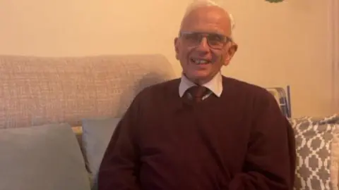 BBC A man with short white hair wearing glasses is smiling and wearing a burgundy jumper with a burgundy tie and white shirt underneath. He is sat on a grey sofa with three grey pillows on it.